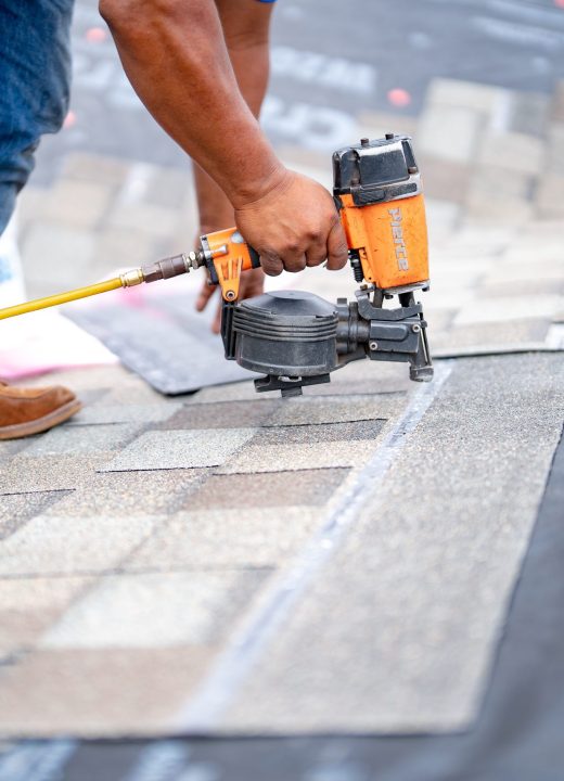 roofing-installation-worker-nailing-shingles-pneumatic-nail-gun-yellow-hose-brown-boots-blue-jeans-roofing-materials-gray-shingles-roof-repair-construction-worker-roofing-project