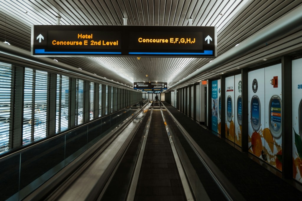 Empty airport walkway with directional signs to hotel and concourses, featuring advertisements on the walls and a smooth walking path.