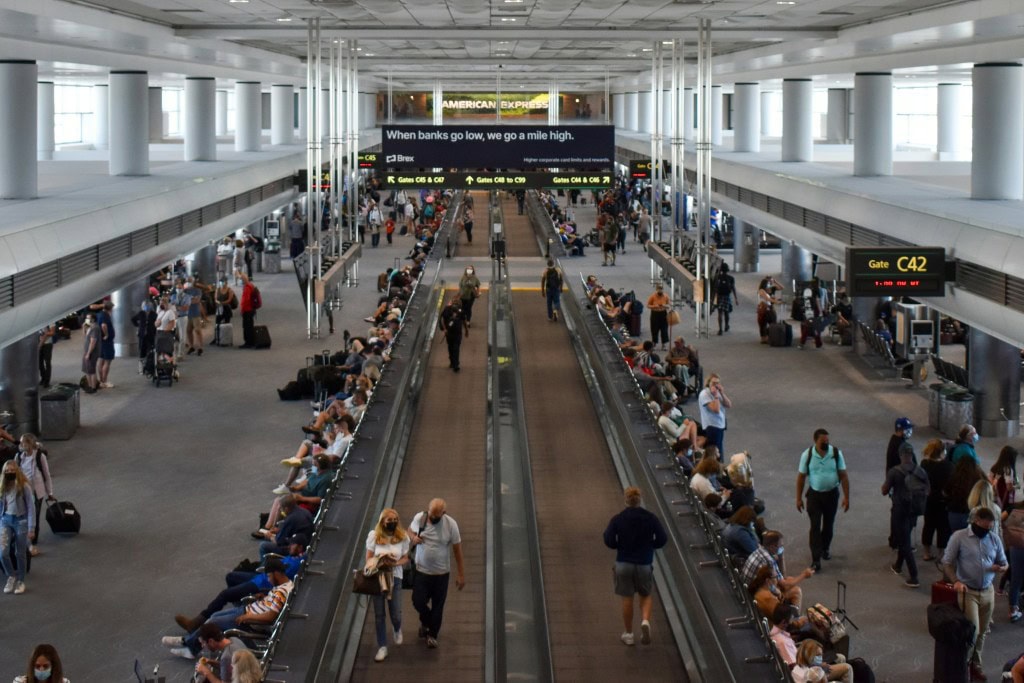 An airport terminal scene with travelers sitting on benches and walking along a moving walkway, with visible gates and signage.