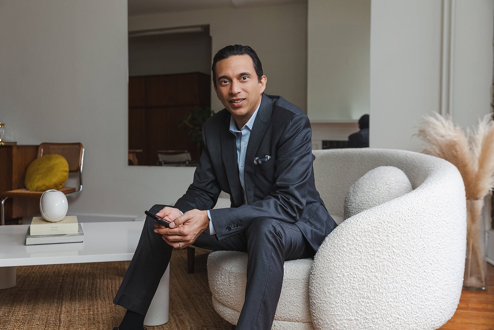 A man in a gray suit sits on a modern white chair holding a smartphone, in a stylish living room with neutral decor—perhaps researching whole life insurance and policy loans as part of his infinite banking strategy.