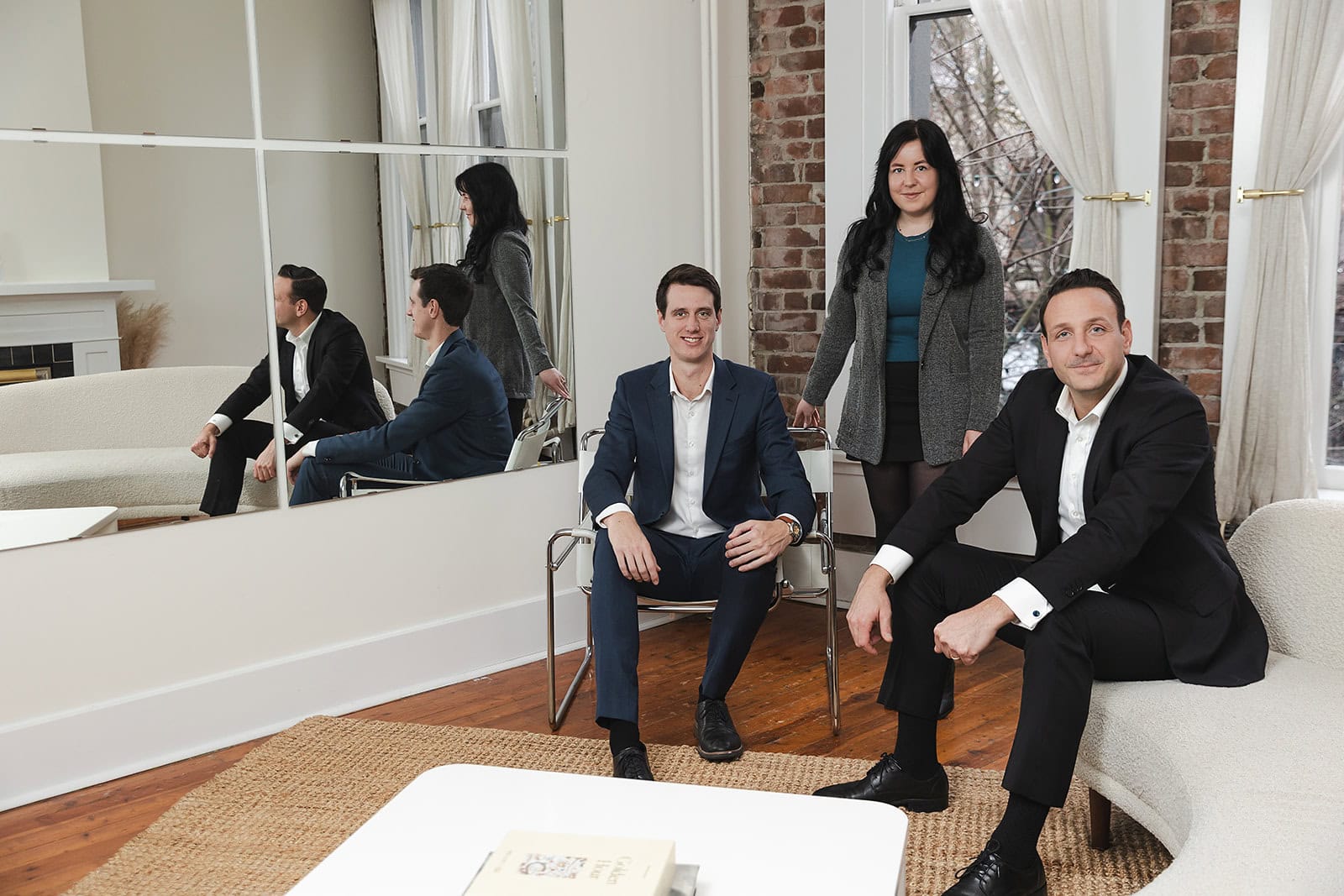 Three professionally dressed people pose in a stylish, modern living room with brick accents and white furniture, confidently discussing whole life insurance. Two men sit while a woman stands behind them, all smiling at the camera.
