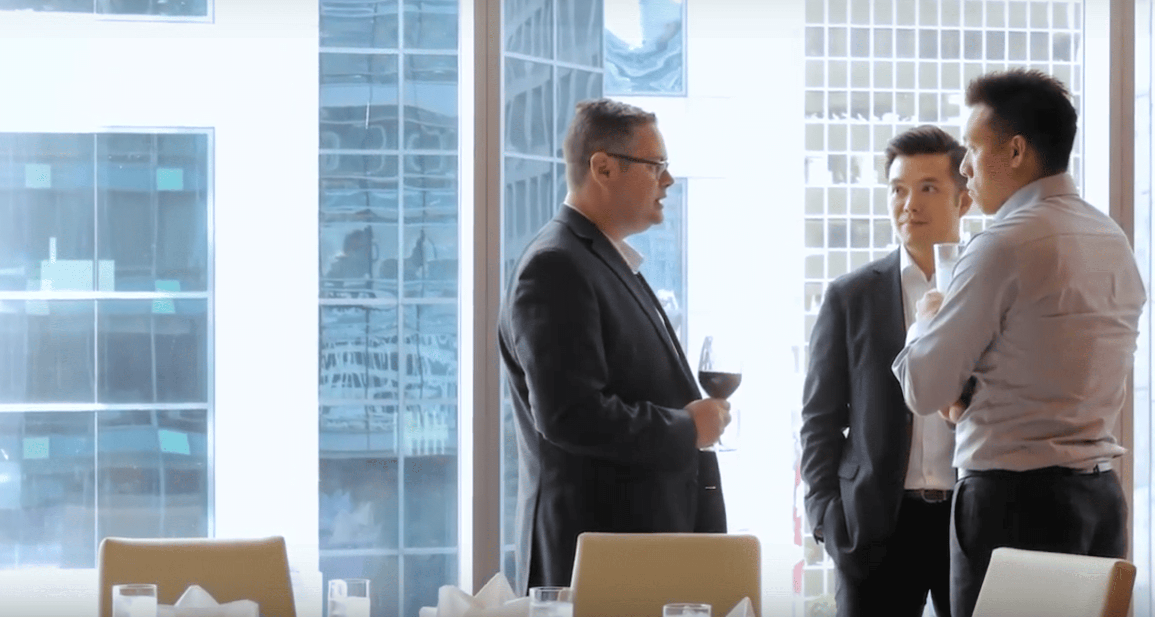 Three men in business attire discuss using life insurance to fund major purchases in a modern, bright office with large windows overlooking city buildings.