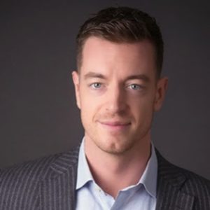 A wealth advisor with short brown hair, dressed in a sophisticated pinstriped suit jacket and light blue shirt, smiles subtly while standing before a dark backdrop—reflecting Safe Pacific Financial’s commitment to bespoke Canadian life insurance policies, personalized wealth management solutions, expert financial advice for affluent families, and the innovative infinite banking strategy.