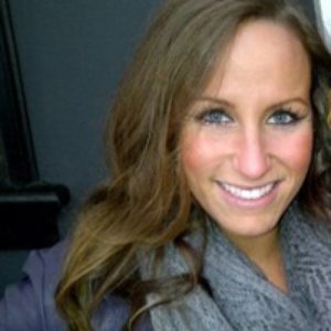 A confident woman with long, wavy brown hair smiles warmly at the camera, embodying the trusted expertise of Safe Pacific Financial. She is dressed in a gray scarf and a dark jacket, reflecting the professionalism associated with wealth management and bespoke Canadian life insurance solutions. The softly blurred dark background emphasizes her approachable demeanor, representing personalized financial advice for affluent families and advocates of the infinite banking strategy.
