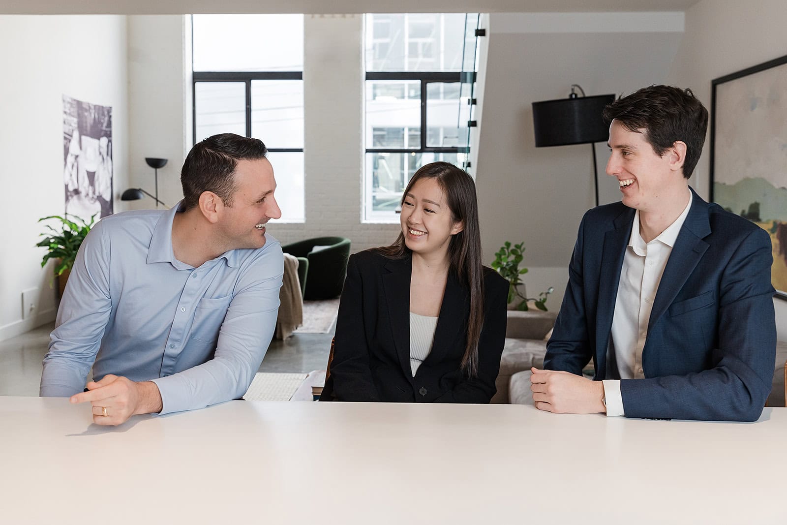 Three professionally dressed people sit at a white table in a modern, bright office, smiling and having a friendly conversation.