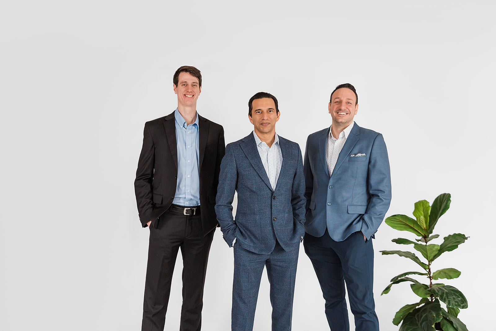 Three men in suits stand side by side against a plain white background, smiling at the camera. There is a green potted plant to the right of the group.