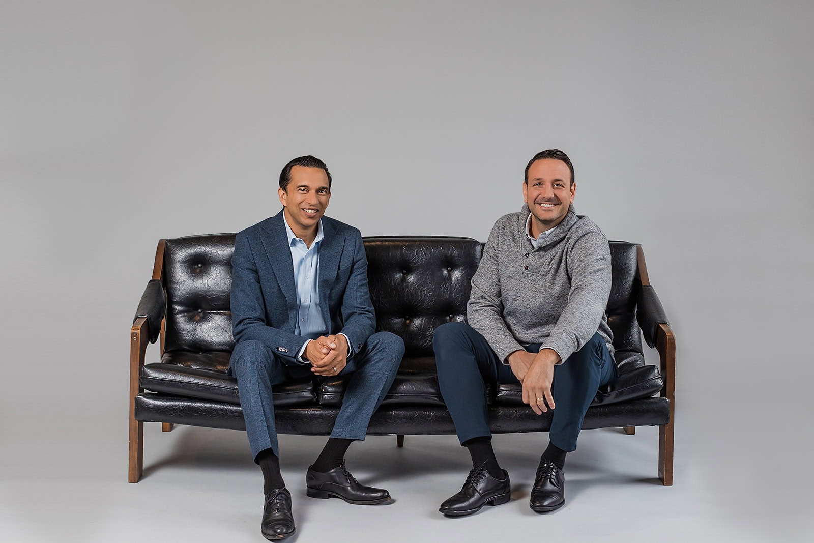 Two men sit on a black leather couch against a plain grey background. One wears a blue suit and black shoes, while the other wears a grey sweater, blue pants, and black shoes. Both are smiling and looking at the camera.