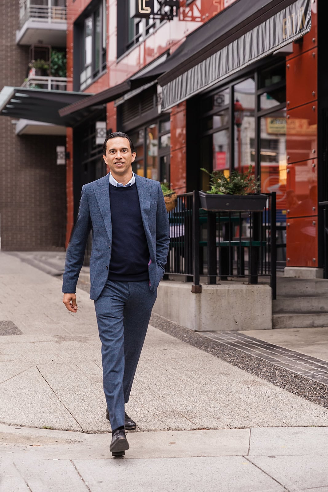 A man in a blue suit with a navy sweater walks confidently down a city sidewalk, with modern buildings and a restaurant patio in the background.