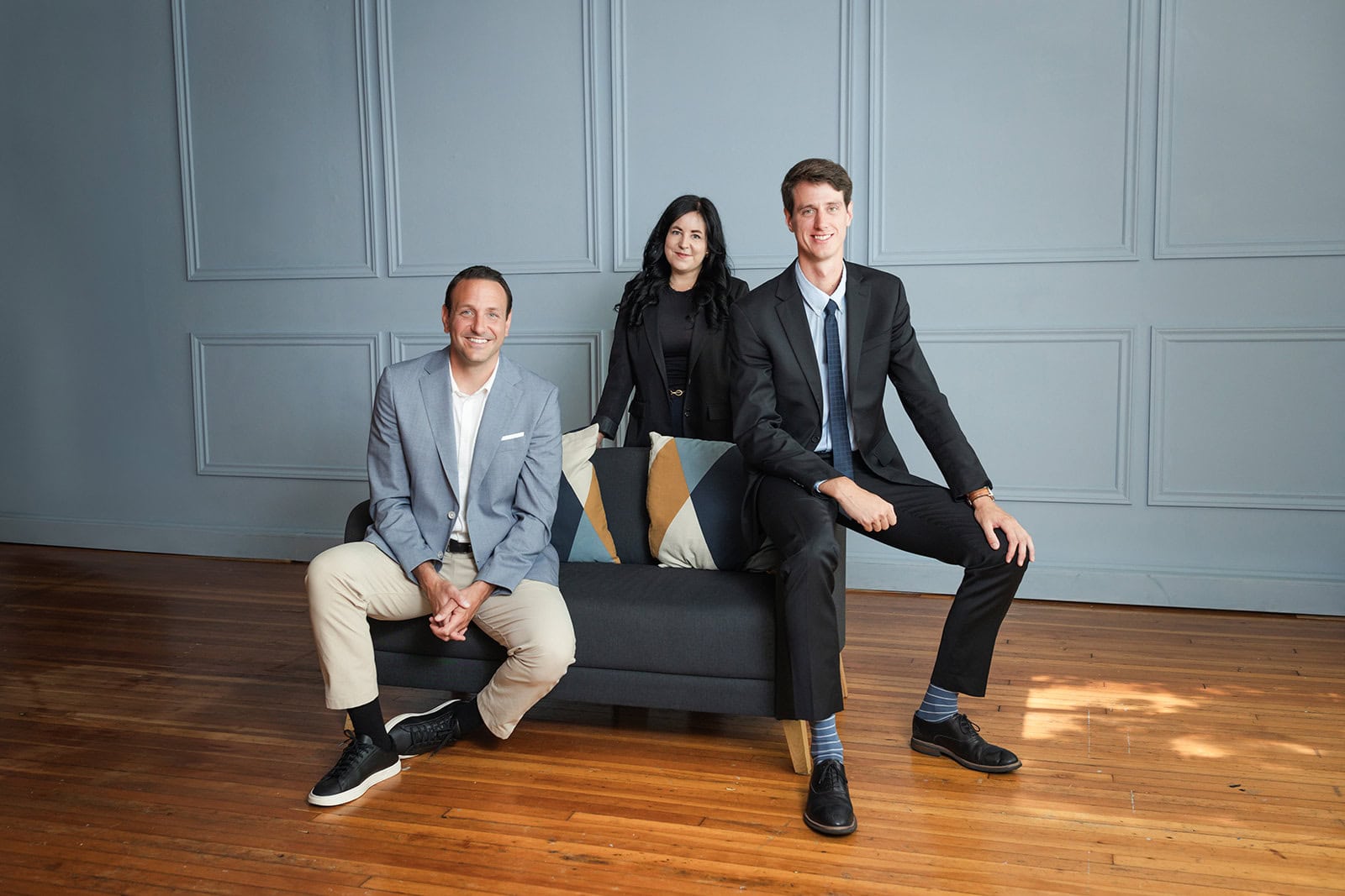 Three people in business attire pose in a modern room with wooden floors and a gray paneled wall. Two men sit on a dark sofa with geometric pillows, while a woman stands behind them, all smiling at the camera.