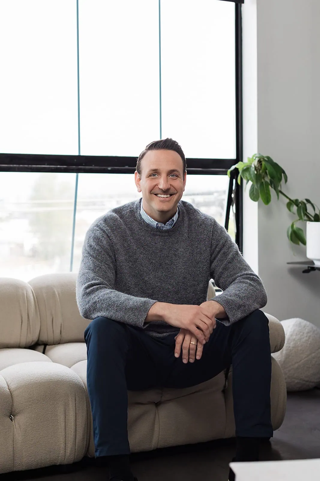 A man in a gray sweater and blue pants sits smiling on a beige sofa in a modern, bright room with large windows and a potted plant in the background.