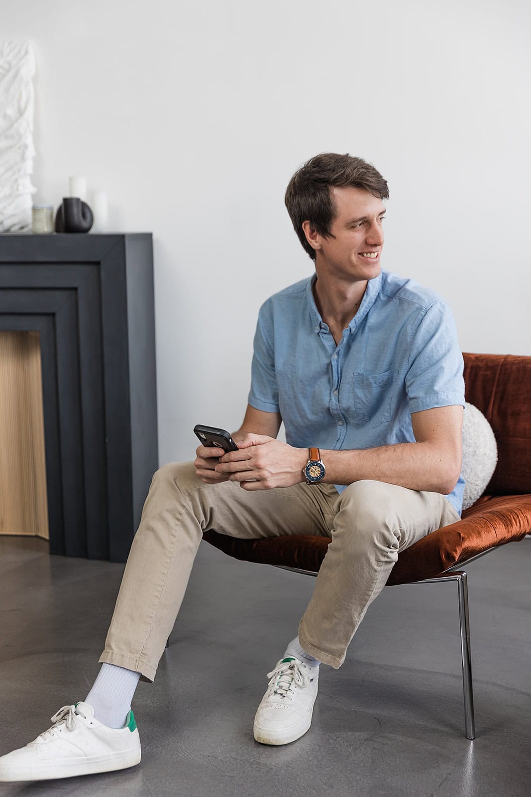 A young man in a light blue shirt and beige pants sits on a brown chair, holding a smartphone and smiling while looking to the side. He wears white sneakers and a watch, with a modern black mantel in the background.