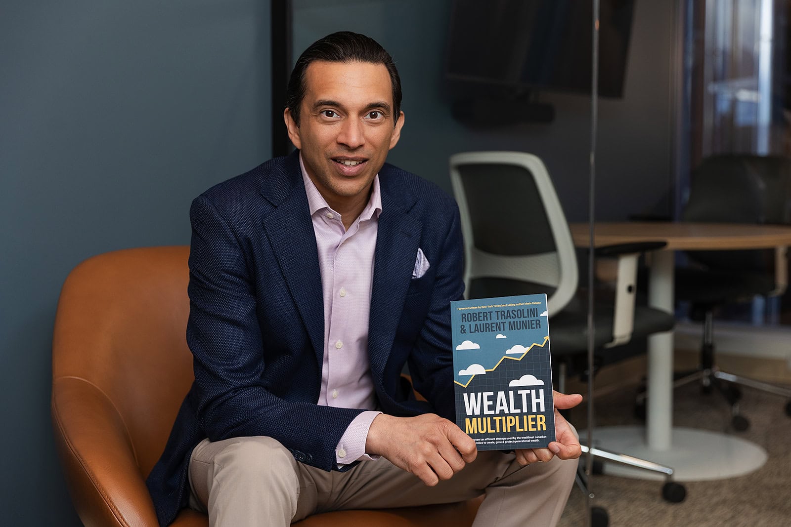 A man in a blue blazer and light pants sits in a modern office, holding Wealth Multiplier, showcasing Safe Pacific Financial’s bespoke Canadian life insurance, wealth management, and infinite banking strategies.
