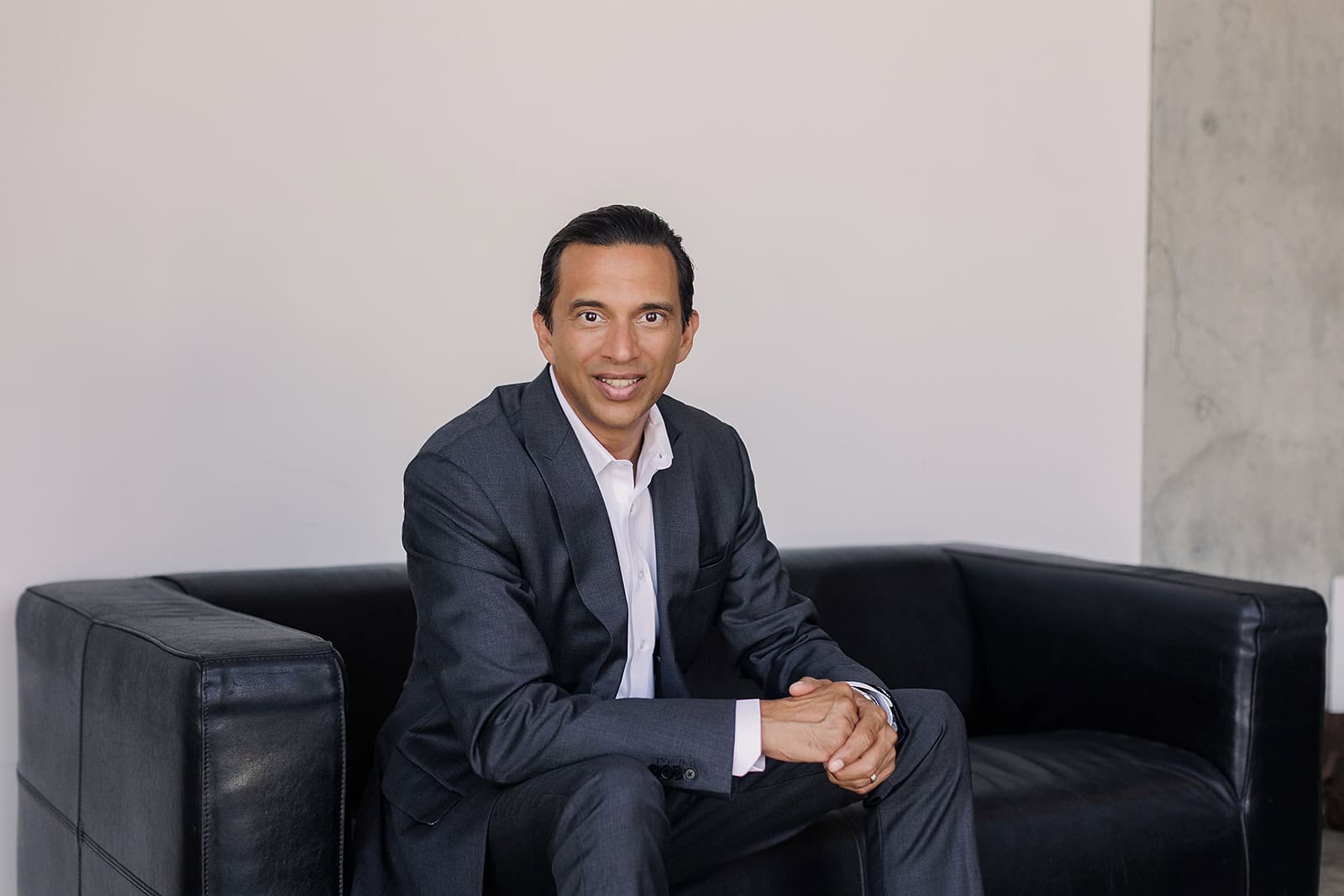A man in a suit and dress shirt sitting on a black leather sofa for a Safe Pacific Financial Discovery VSL booking, specializing in wealth management, the infinite banking strategy, and bespoke Canadian life insurance policies for affluent families.