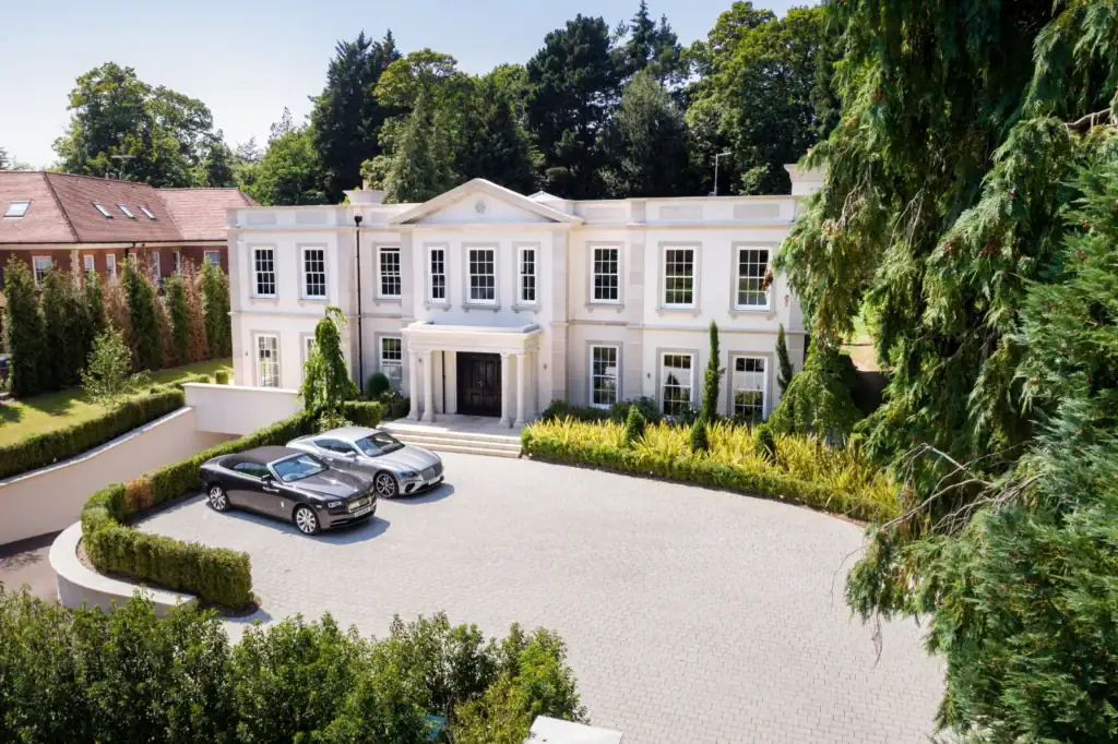 A large, white, two-storey mansion with columns and many windows features smart home automation and cinema installation. Two luxury cars are parked on its stone drive, surrounded by manicured greenery and trees in Cobham, Surrey.