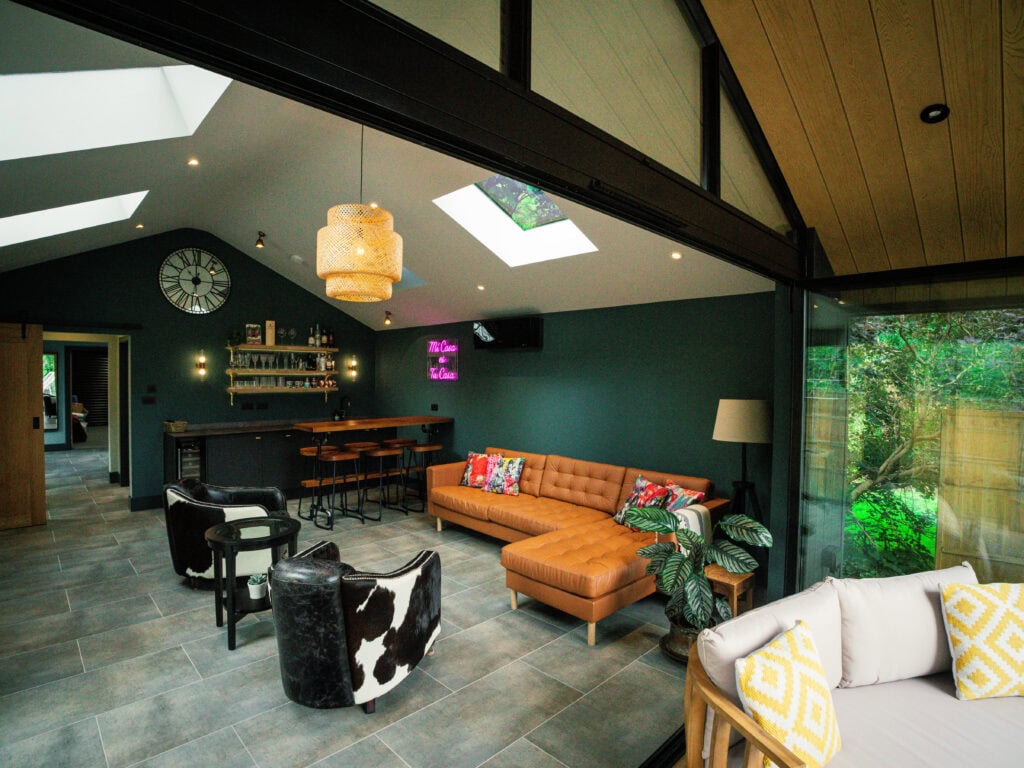 Garden room lounge with a tan leather corner sofa, cowhide armchairs, and a bar with stools and open shelving under a vaulted ceiling with skylights