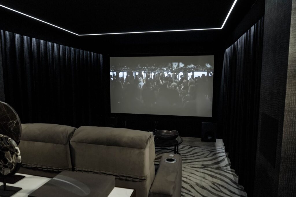 Home cinema room with dark walls, a large projector screen showing a film scene and a grey sofa