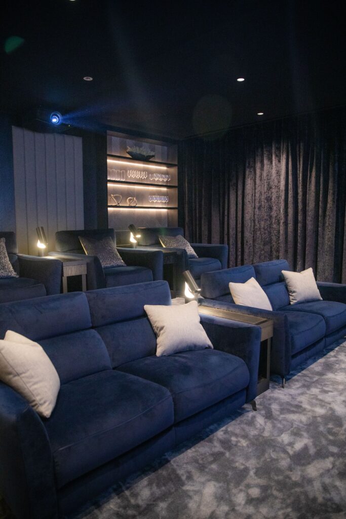 Two rows of navy blue recliner seats in a media room under soft lighting, with side tables and lit shelves of glassware against a dark wall