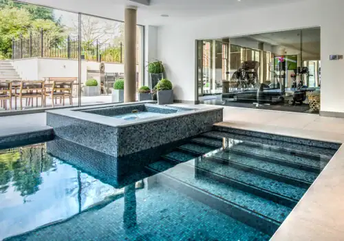 Indoor pool with built-in hot tub, tiled steps, large glass doors to a patio with outdoor dining, and a custom-built gym with cinema in Weybridge Surrey visible through another glass wall. Modern, bright design enhanced by potted plants.