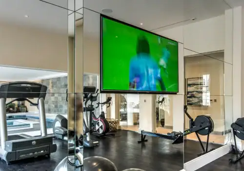 A modern home gym with a treadmill, rowing machine, exercise bike, and large wall mirror. Enjoy your workout while a big TV—perfect for cinema installation in Weybridge Surrey—displays a sports match, with a swimming pool visible through the window.