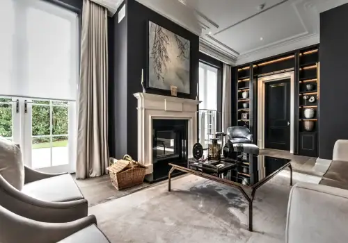 A modern living room with dark walls, a fireplace, built-in shelves with decor, and smart lighting installation in Oxshott Surrey. Light chairs on a plush rug surround a glass coffee table, while a woven basket sits near the fireplace.