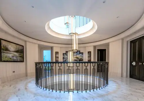 A modern, circular corridor with marble floors, a central spiral staircase with glass balustrade, a large round skylight, elegant home automation lighting and shading in Oxshott Surrey, black double doors, and landscape paintings on the walls.