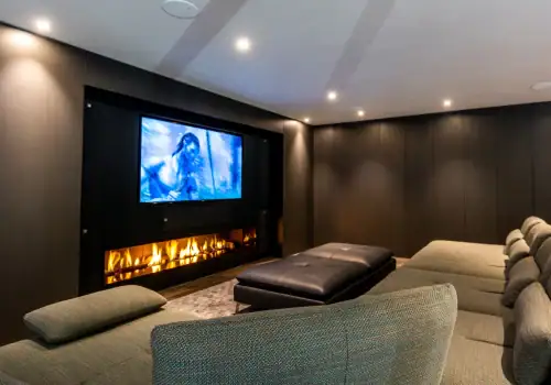 A modern home theatre room featuring a home cinema installation in Esher Surrey with a wall-mounted TV showcasing a film, a cushioned sectional sofa, leather ottoman, sleek linear fireplace beneath the TV, and ambient recessed ceiling lights.
