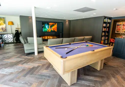 A modern living room with a wooden pool table set, grey sectional sofa, fireplace, bookshelves, and art on the walls features a dark herringbone floor—perfect for relaxing or enjoying your new home cinema installation in Esher Surrey.