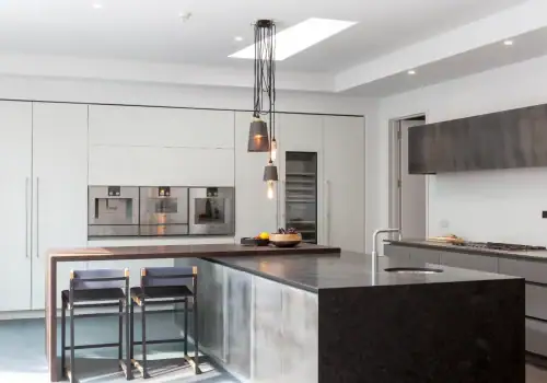 Modern kitchen with a large dark island worktop, two black chairs, built-in stainless steel appliances, minimalist white cabinetry, pendant lights above the island, and seamless home automation installation in Hampstead.
