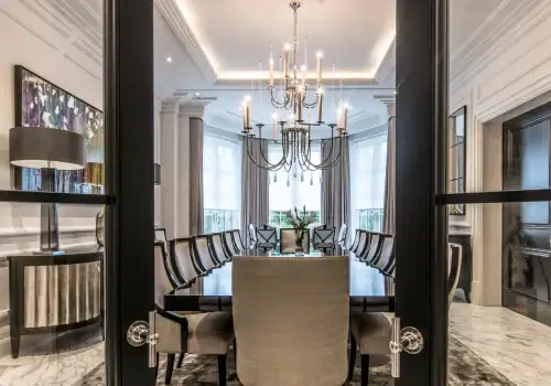A modern dining room with a long table, ten chairs, a large chandelier, tall windows with sheer curtains, elegant decor, and seamless smart lighting installation in Oxshott Surrey for effortless lighting and ambience control.