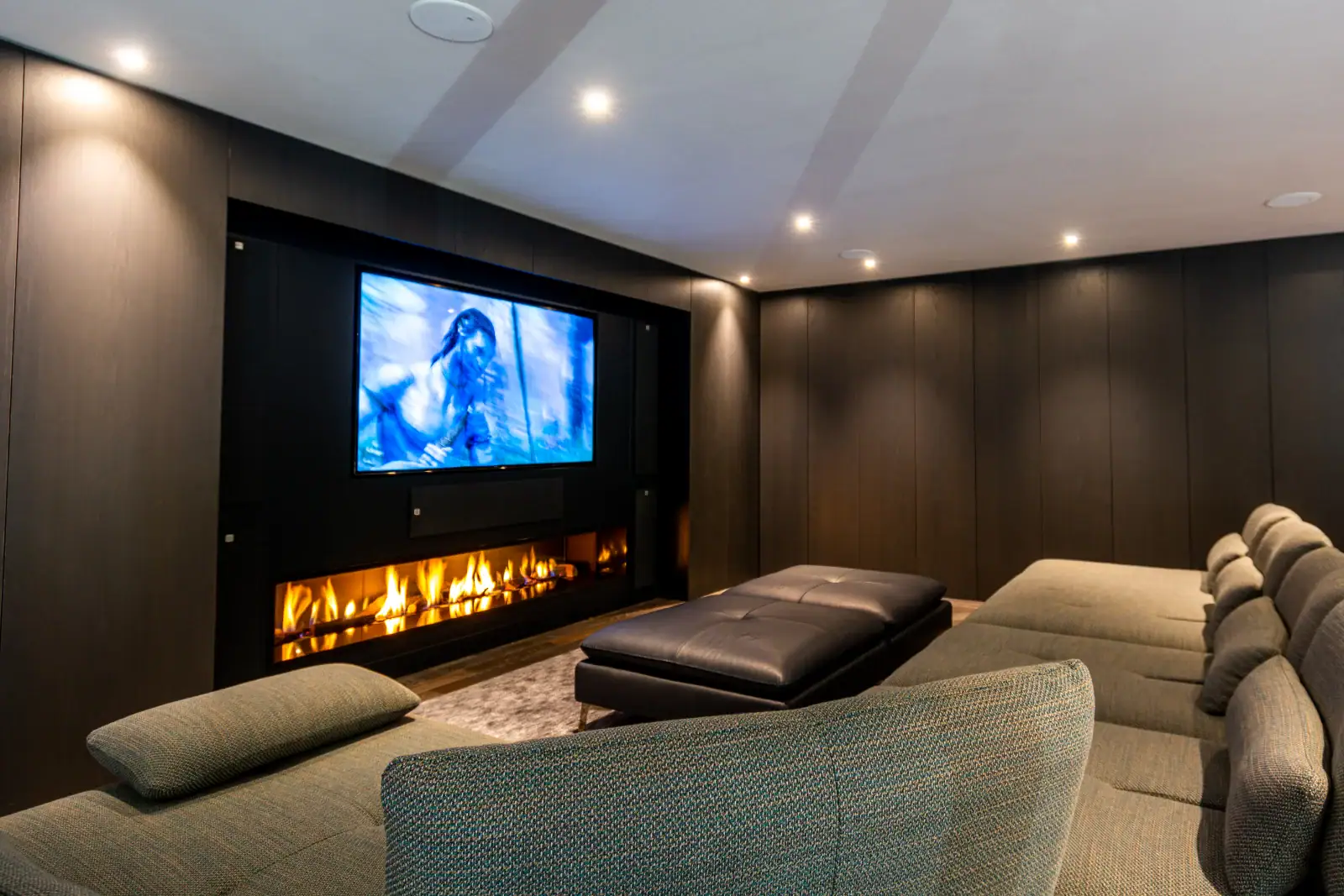 A modern home theatre room with a large wall-mounted TV displaying a film scene, a long sectional sofa, black ottoman, recessed lighting, and a sleek built-in fireplace—perfect for luxury home entertainment and cinema installation in Esher, Surrey.