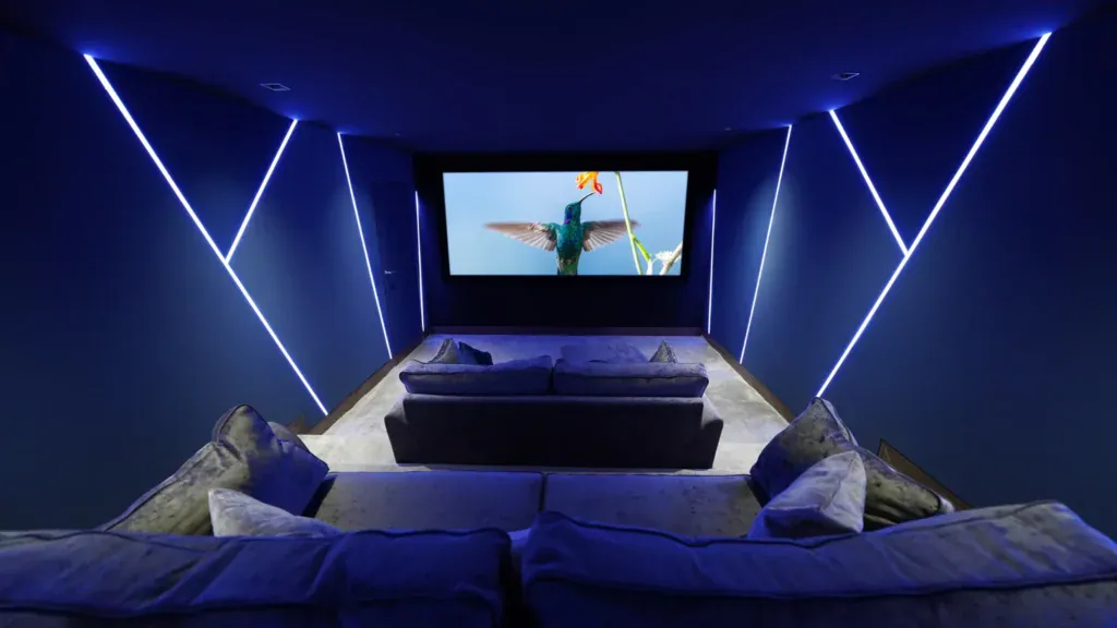 A modern home theatre room with blue LED lighting, plush grey sofas, and a large screen displaying a close-up of a hummingbird in flight