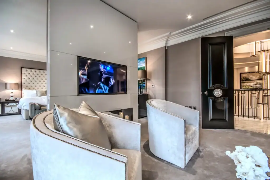Modern luxury bedroom with neutral tones, velvet armchairs, a mounted flat-screen TV—ideal for a home cinema installation in Oxshott, Surrey—plush bedding, and sleek decor. An open black door reveals a lit hallway and elegant staircase railings.