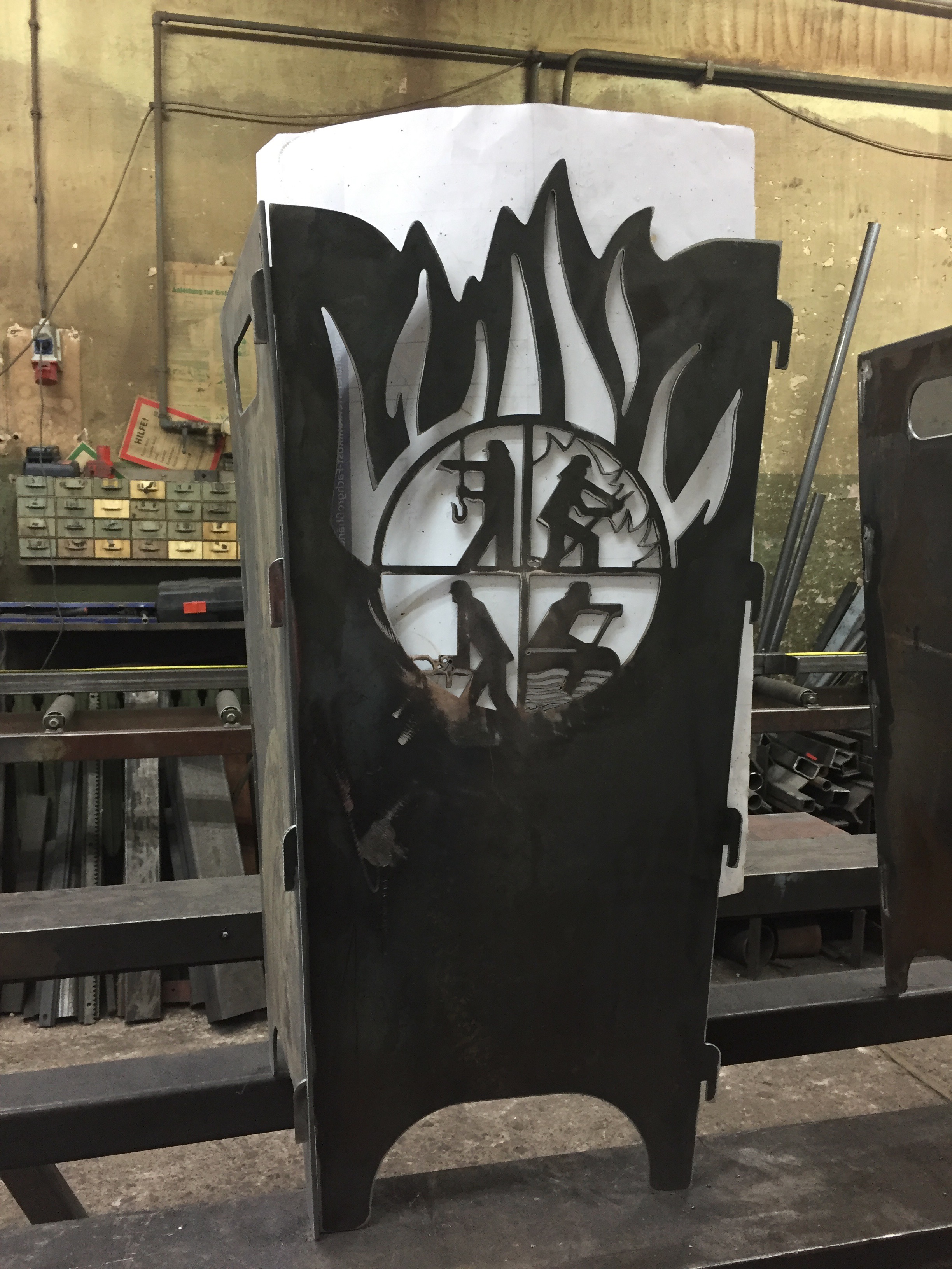 Robuster Metall-Feuerkorb von Sonderanfertigungen von Stahl- und Metallbau Drost mit lasergeschnittenen Flammen- und vier Handwerker-Silhouetten-Motiven.