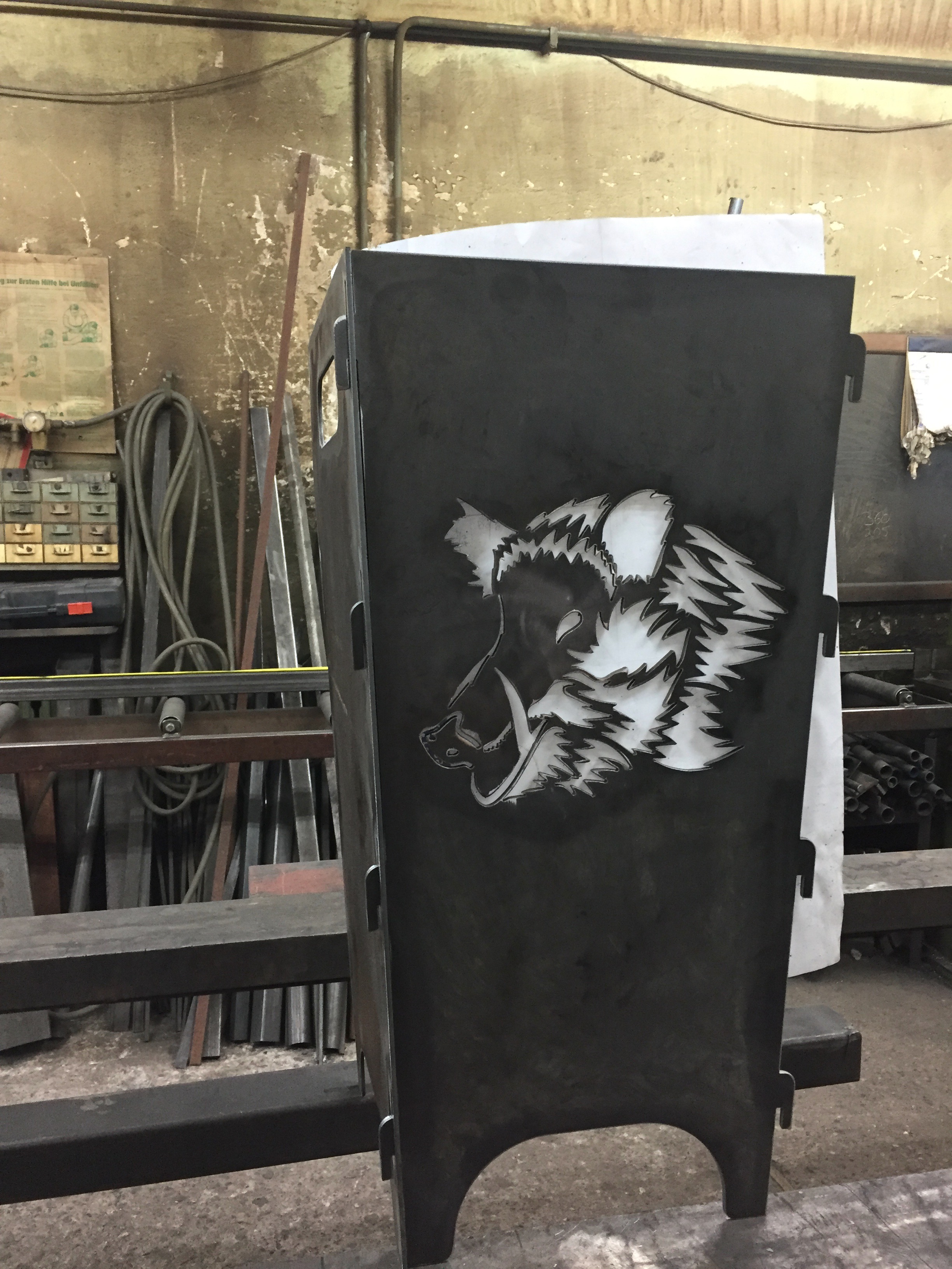 Dunkelgraue Metall-Feuerstelle mit einem prägnanten, lasergeschnittenen Wildschweinkopf-Motiv an der Vorderseite, gefertigt von Sonderanfertigungen von Stahl- und Metallbau Drost.