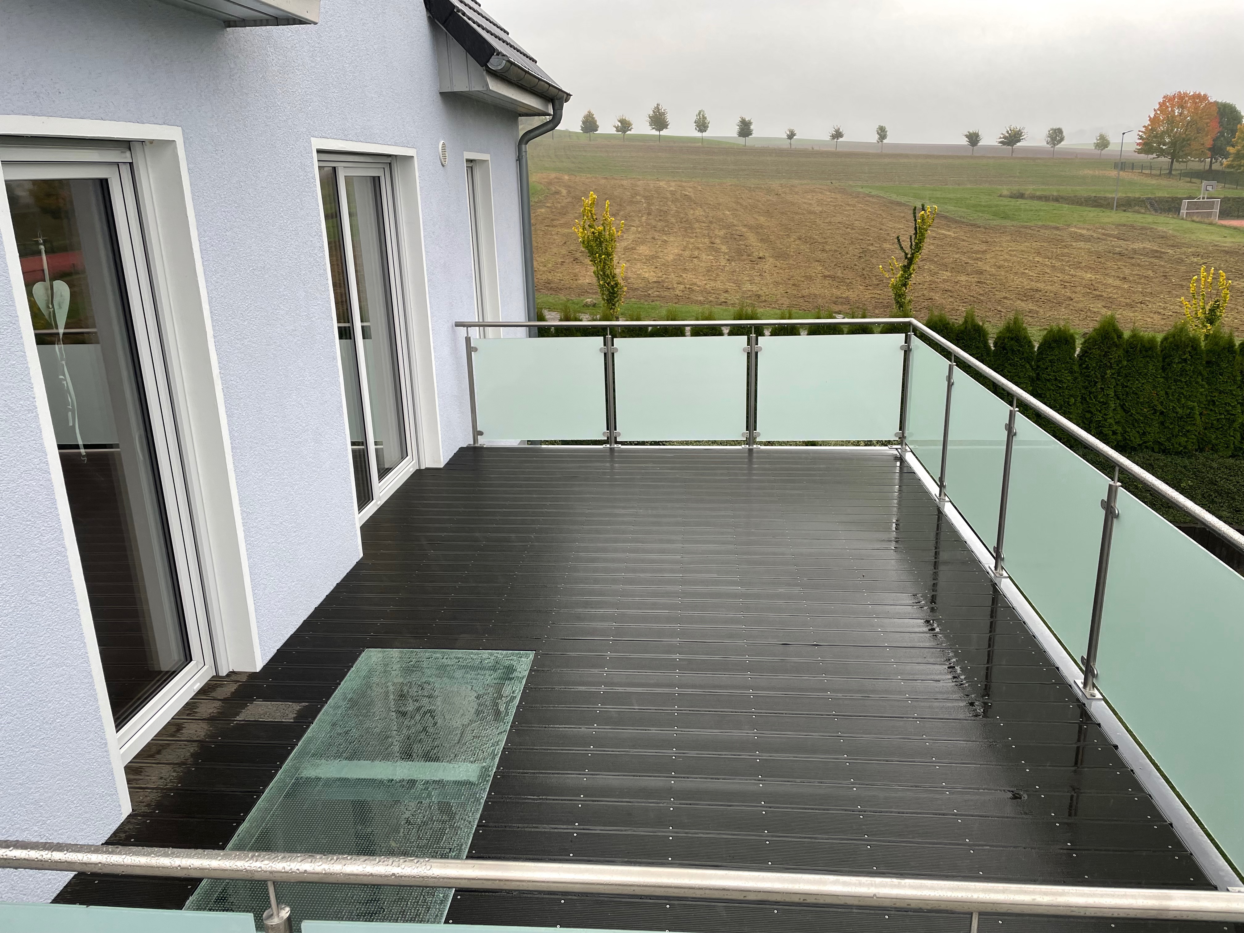 Moderner Balkon von Stahl- und Metallbau Drost mit dunklem Dielenboden, Edelstahlgeländer, Milchglas-Füllungen und einem integrierten Bodenglas-Element, an einem hellblauen Haus.