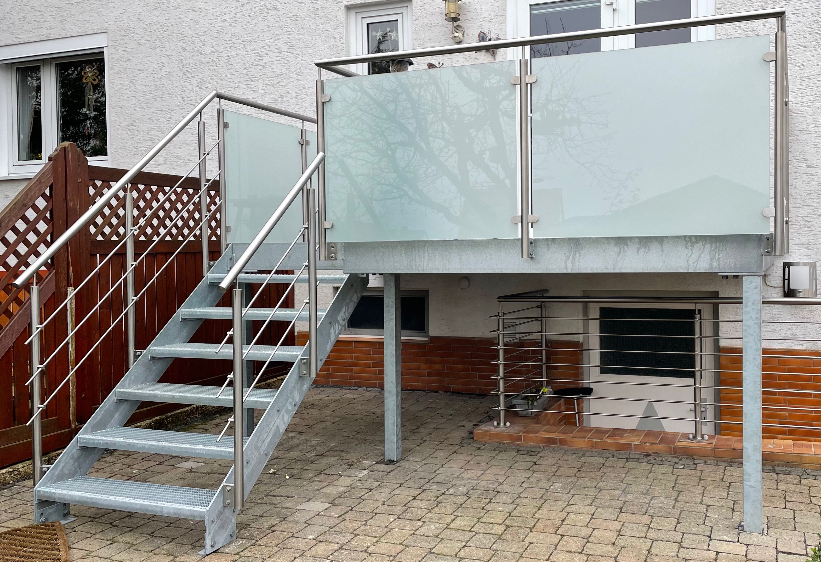Balkon von Stahl- und Metallbau Drost: Freistehender verzinkter Stahlbalkon mit Außentreppe, Edelstahlgeländer und satinierten Glasbrüstungen vor einem Wohnhaus.