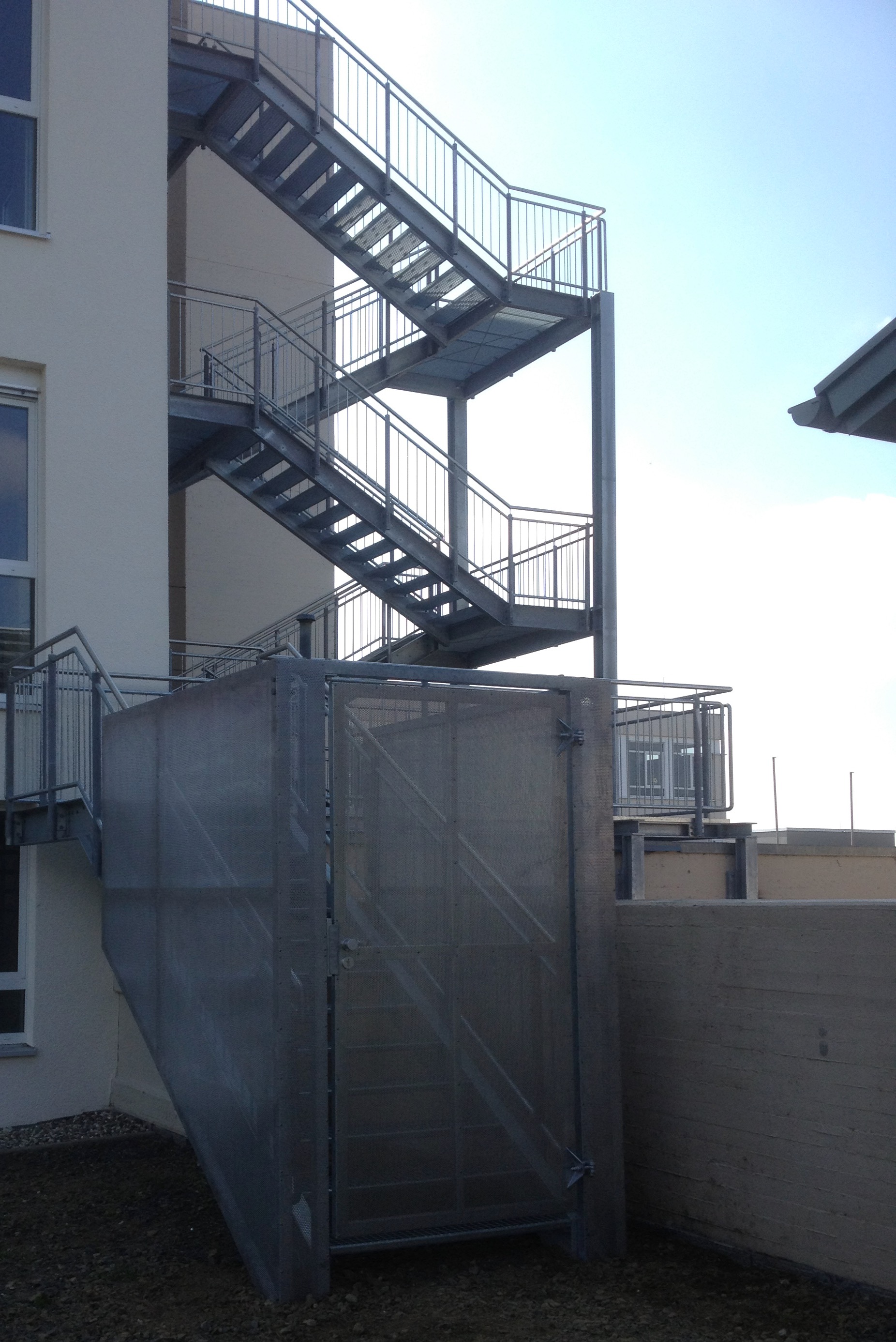 Mehrgeschossige, verzinkte Stahl-Außentreppe mit filigranem Geländer, an einem hellen Gebäude befestigt. Der untere Zugang ist durch ein Metallgitter-Gehäuse gesichert. Eine hochwertige Treppe von Stahl- und Metallbau Drost.