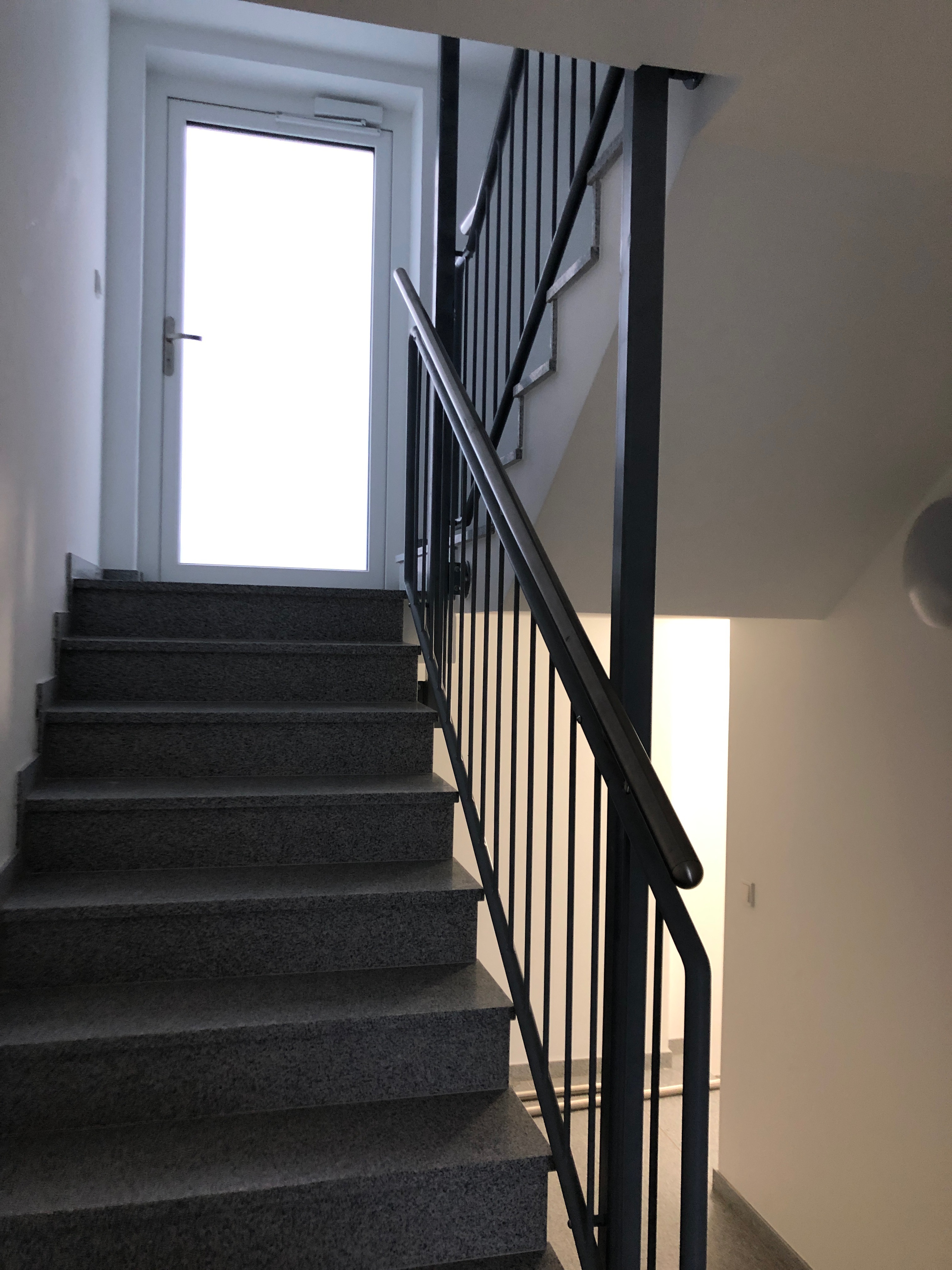 Modernes, gerades Treppengeländer von Stahl- und Metallbau Drost aus dunklem Metall mit vertikalen Stäben und passendem Handlauf, installiert an einer dunkelgrauen Granittreppe in einem hellen Treppenhaus.