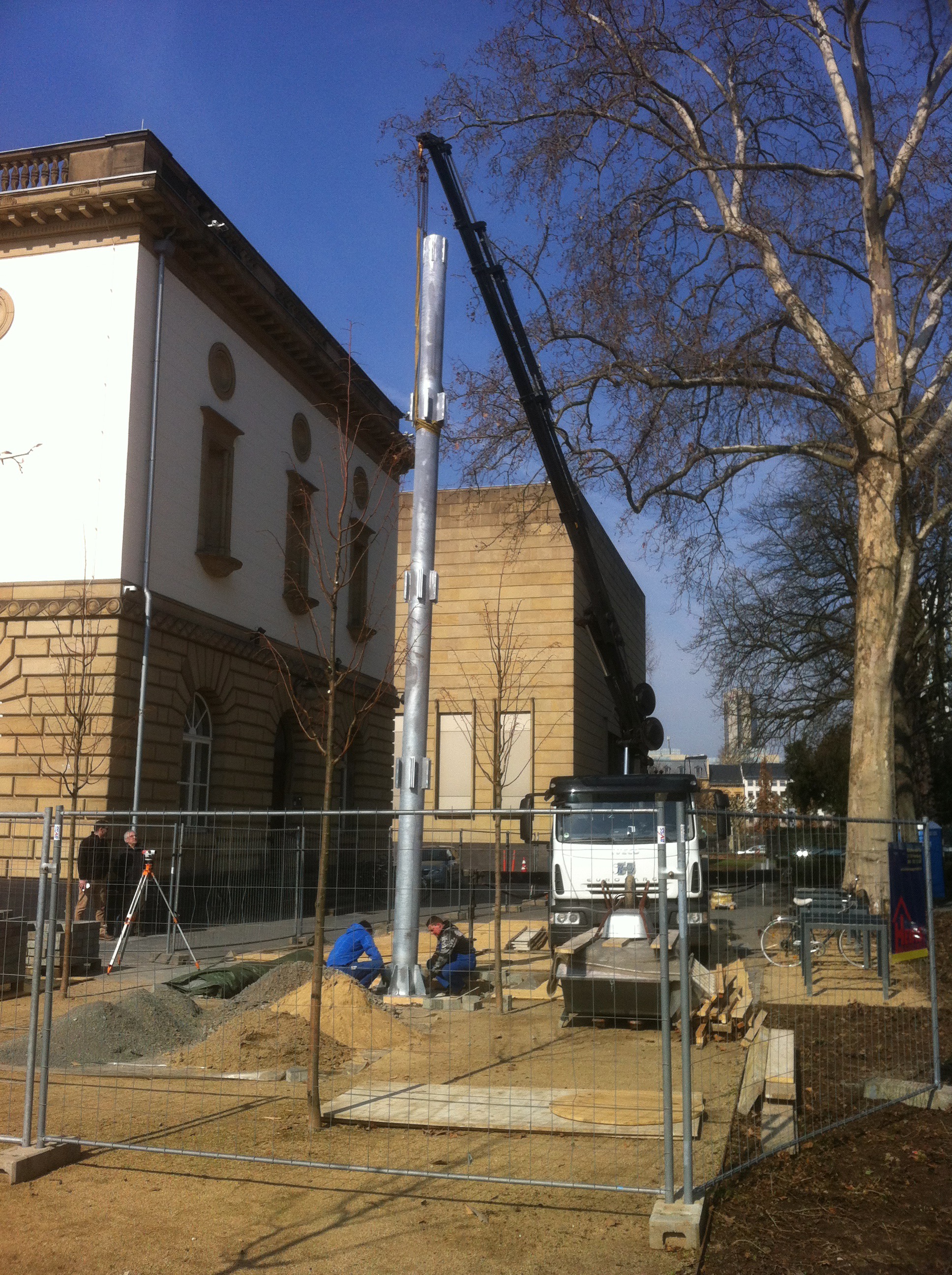 Sonderanfertigungen von Stahl- und Metallbau Drost: Ein Kran installiert einen hohen silbernen Metallmast auf einer Baustelle. Arbeiter sind bei der Montage und Vermessung vor historischen Gebäuden tätig.