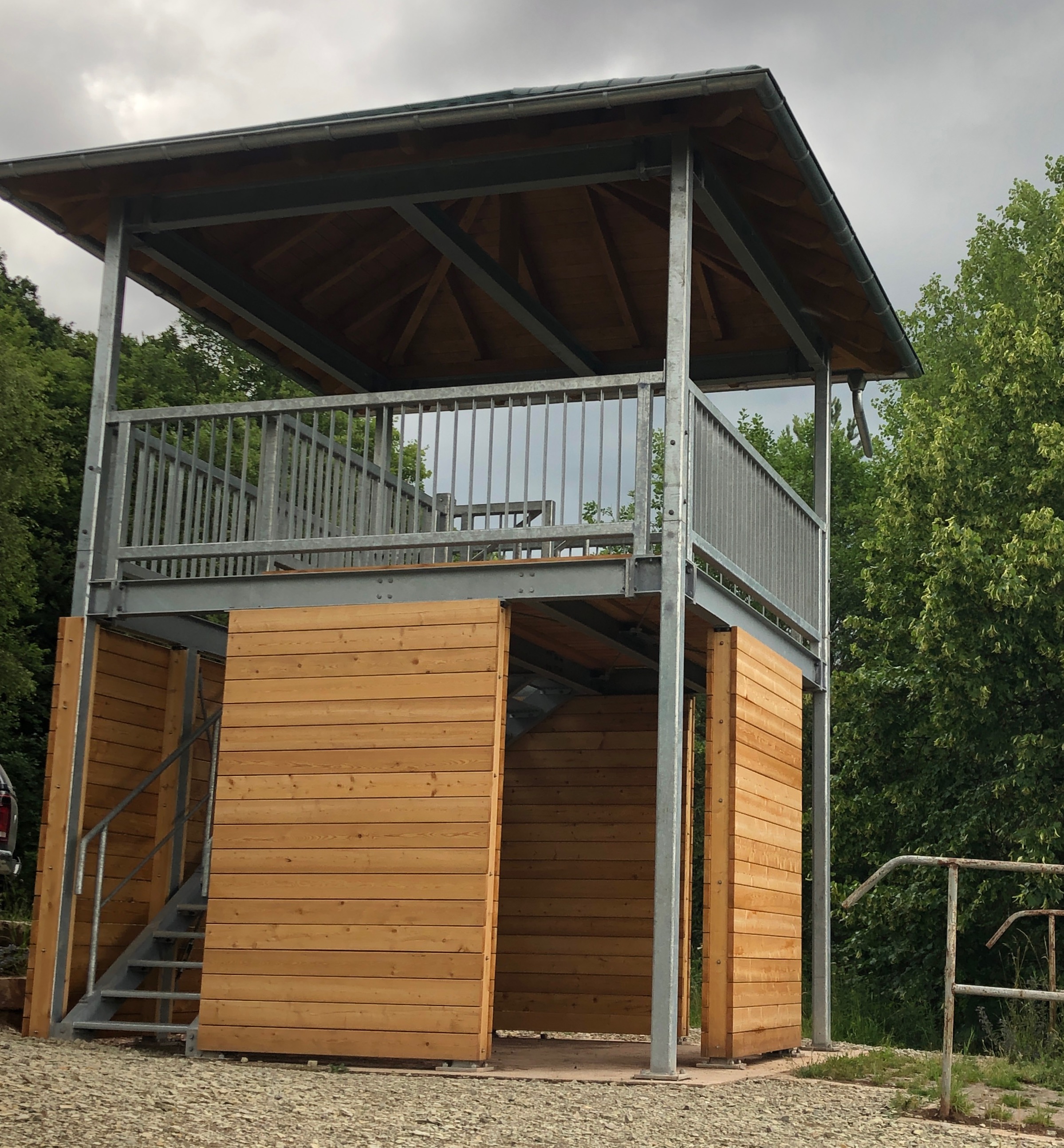 Zweistöckige Stahlkonstruktion von Stahlbau von Stahl- und Metallbau Drost mit Holzverkleidung im unteren Bereich, offener oberer Etage mit Metallgeländer und schrägem Holzdach, umgeben von Bäumen.