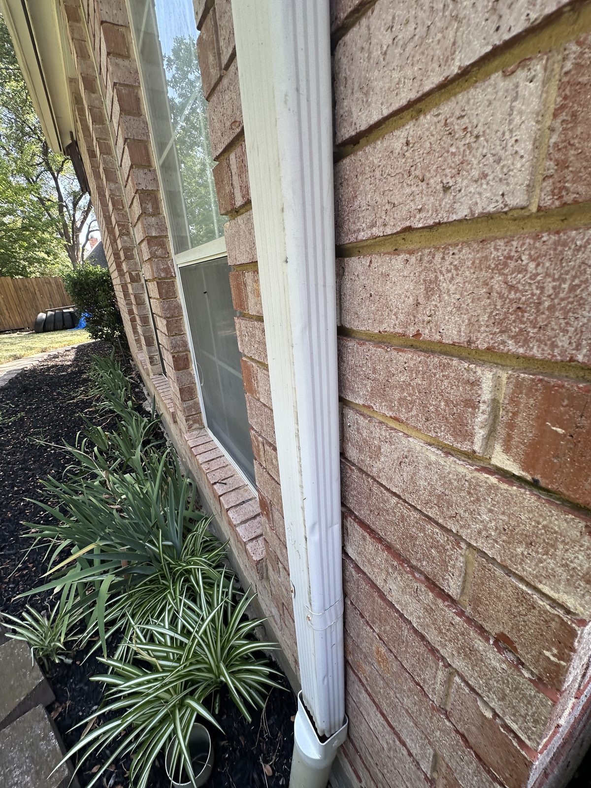 White downspout next to brick wall with landscaping visible