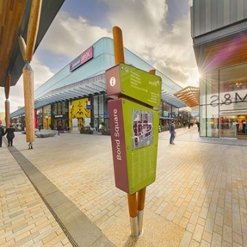 illuminated wayfinding totem signs designed for The Lexicon, Bracknell. The sign is made from two wooden poles that are angled to create a V-shape. These enclose two lozenge shaped signs, in lime green with white letters. The larger bottom sign features a map and list of retailers and amenities. The smaller top one provides directional information to selected locations