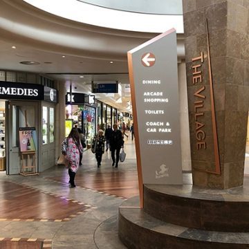 Large totem sign at the entrance to The Village Arcade. The sign is faced in a grey beige colour with a coppery coloured chamfered sides. The content is in white with a copper coloured disc surrounding the directional arrow.