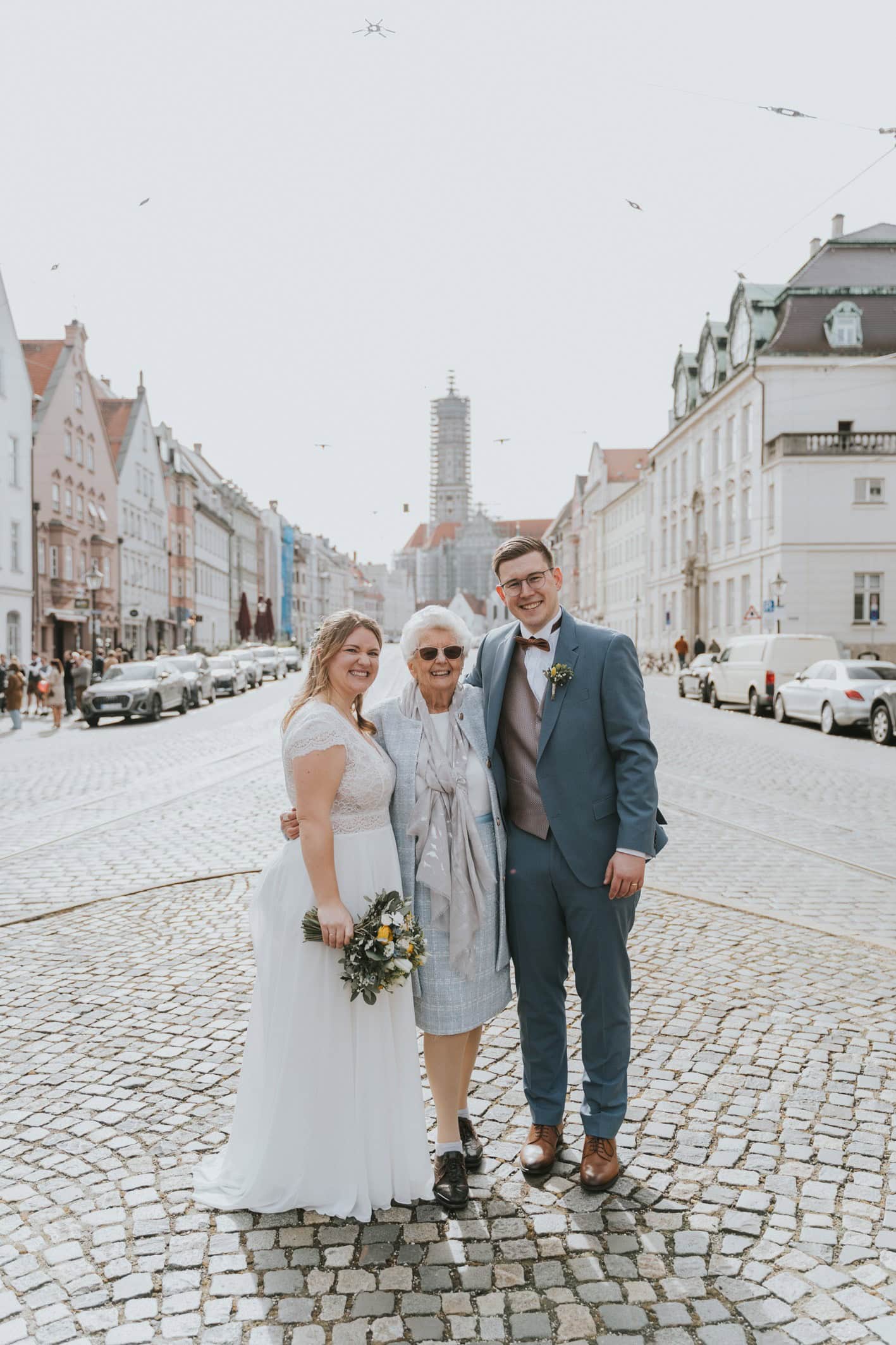 Fröhlicher Moment: Brautpaar und Großmutter lachen – Authentische Hochzeitsfotografie in Augsburg in der Maxstraße