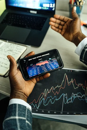 pexels-photo-5583964-5583964 Close-up of hands holding a smartphone with stock market data, charts on paper and laptop in view.