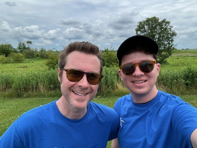 Two men smiling outdoors in matching blue shirts.
