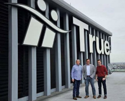 Three men posing near True company sign.
