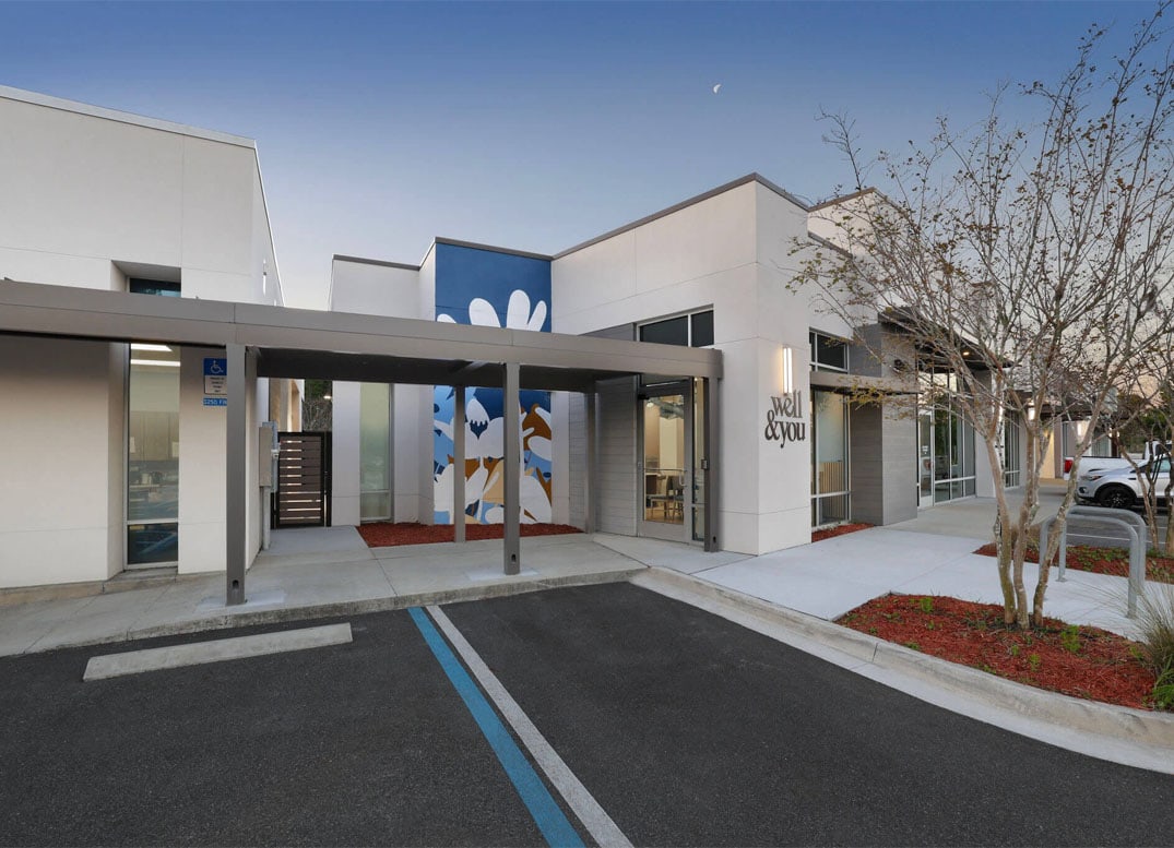 Modern one-story building with white exterior, large windows, a covered entryway, accessible parking, and a blue and white mural on the wall. Small landscaped areas with mulch and trees.