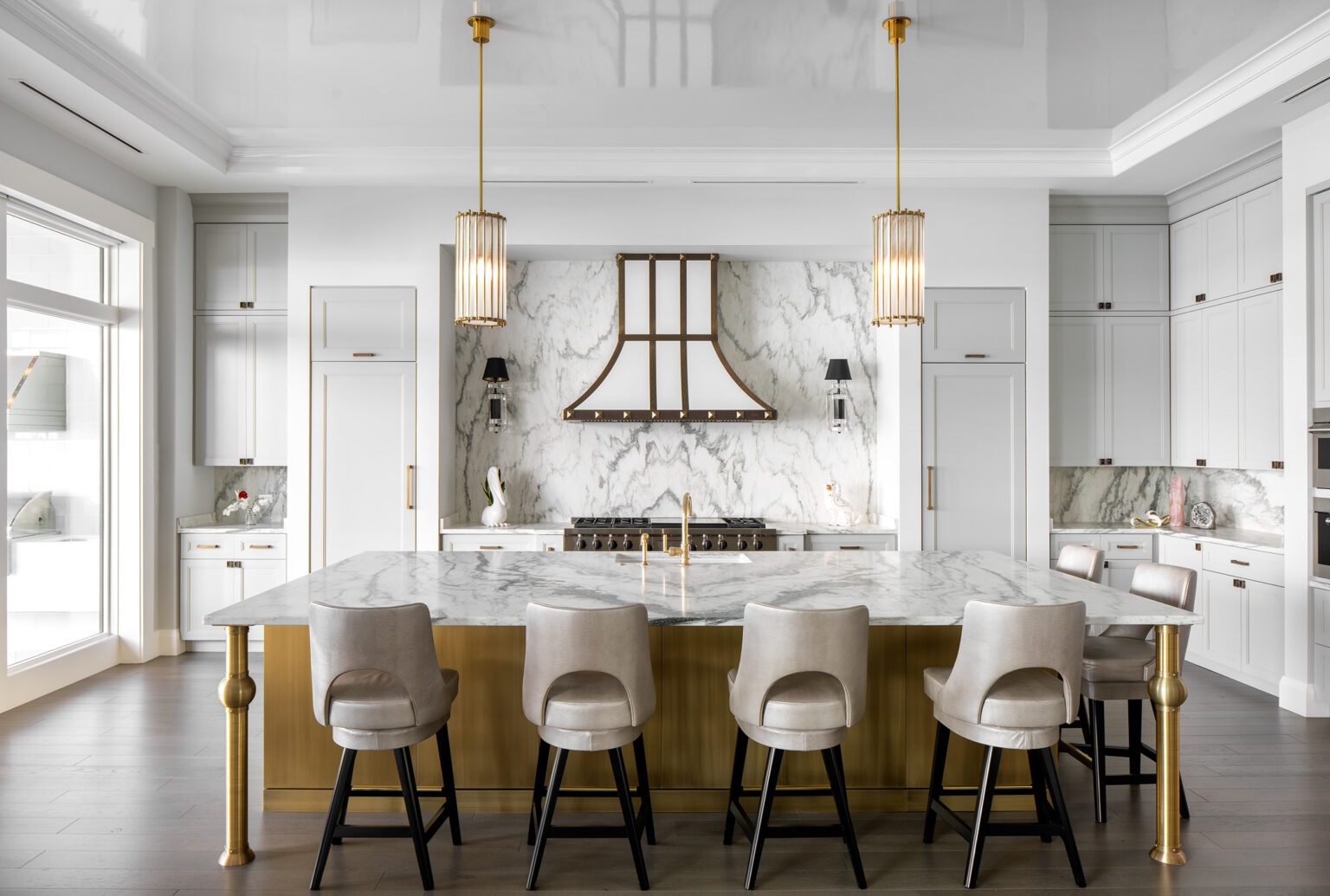 White kitchen with a marble island, brass-trimmed range hood, crystal pendants and upholstered counter stools