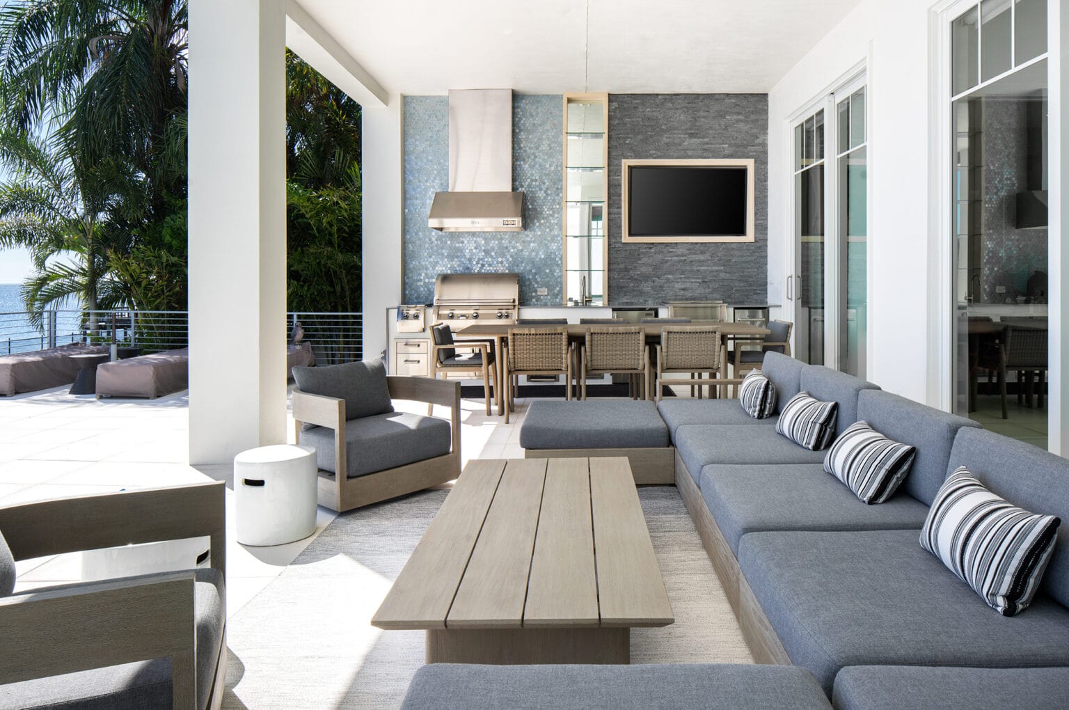 Covered outdoor living room with a gray sectional, a long wood coffee table, an outdoor kitchen and palm trees beyond